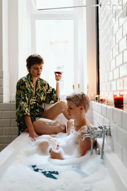 two people sit in a bathtub with bubbles and wine in the tub