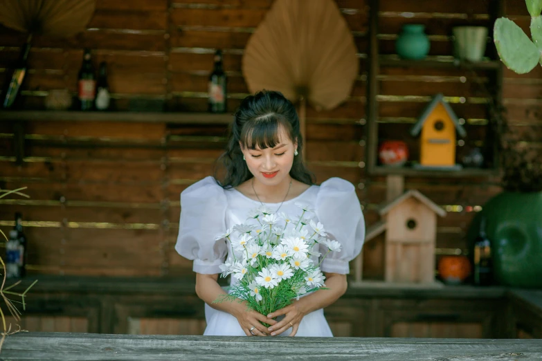 a young woman with long hair standing behind a table holding a bouquet of daisies