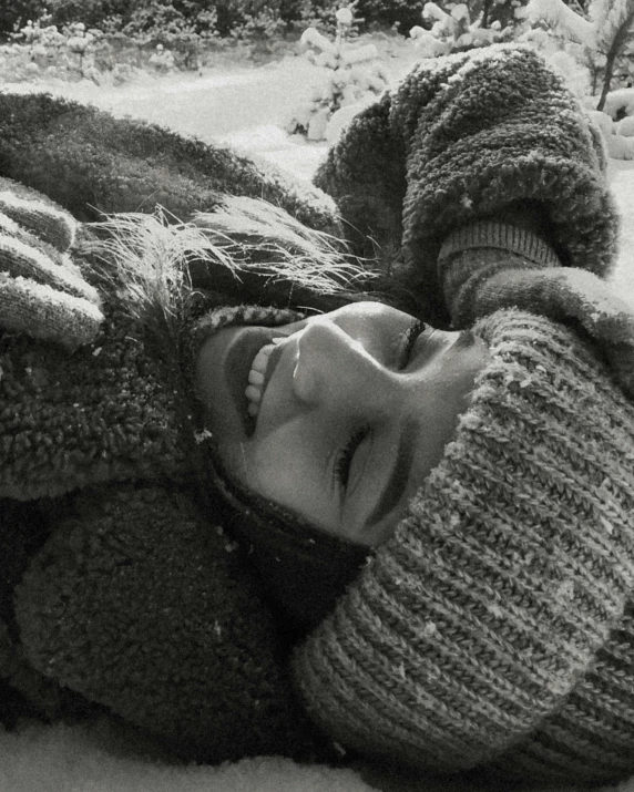 the black and white image shows a young woman laying in the snow in her winter outfit