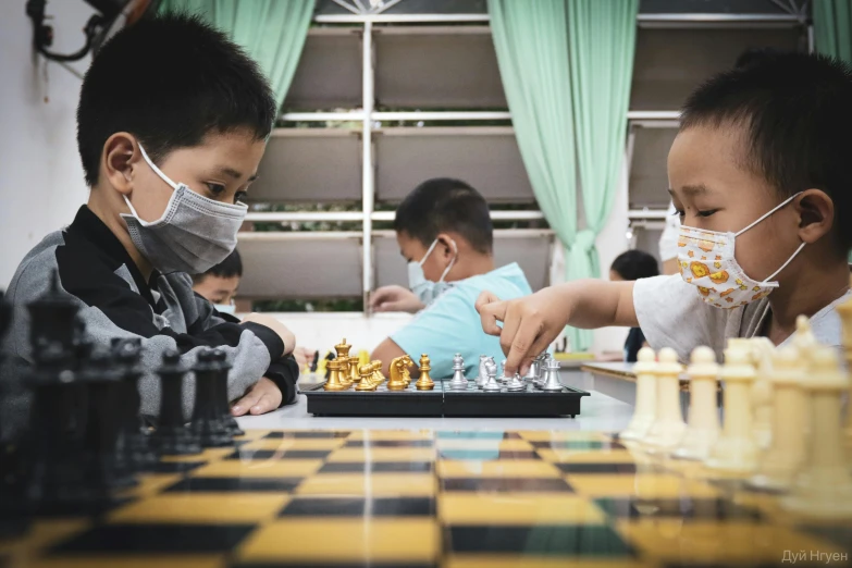 two boys playing chess wearing face masks while a third plays