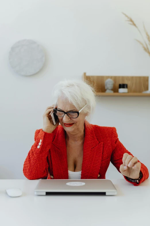 a woman in a red blazer holds her phone up to her ear as she sits in front of a laptop