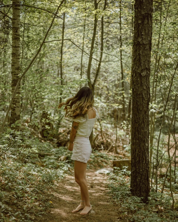 a woman in a short dress stands by trees and looks up at the sun