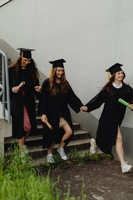 three women in graduation caps and gowns hold hands as they walk down stairs