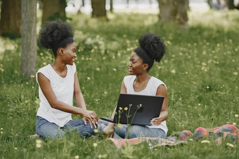 two women sit on the grass with one on the laptop