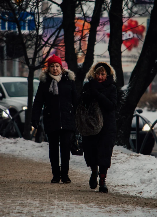 two women walk in the snow past parked cars