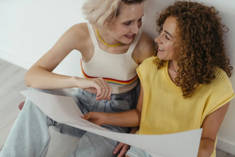 two women sitting against a white wall and looking at soing