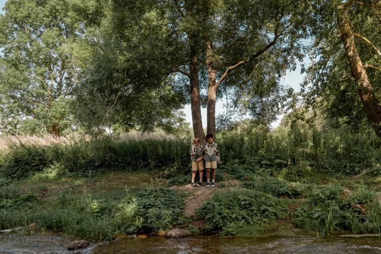 two people standing in the shade of a large tree next to a river
