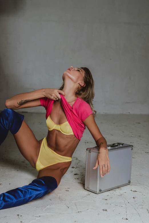 a young woman in a yellow bikini top and blue pants is sitting on the floor, leaning against a suitcase