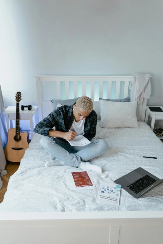 a young man sitting on his bed with an open laptop and a guitar