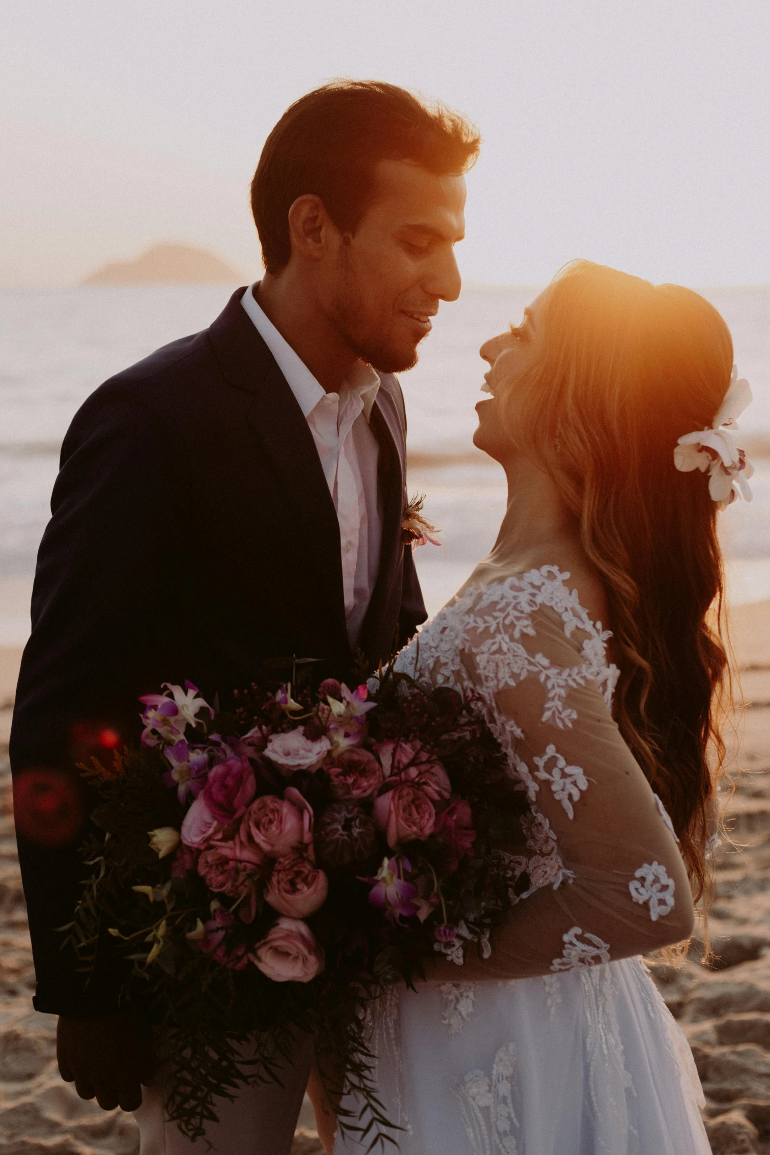 a bride and groom stand together on the beach at sunset