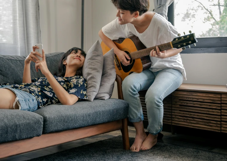 woman is playing the guitar while another person lies on the couch and plays the guitar