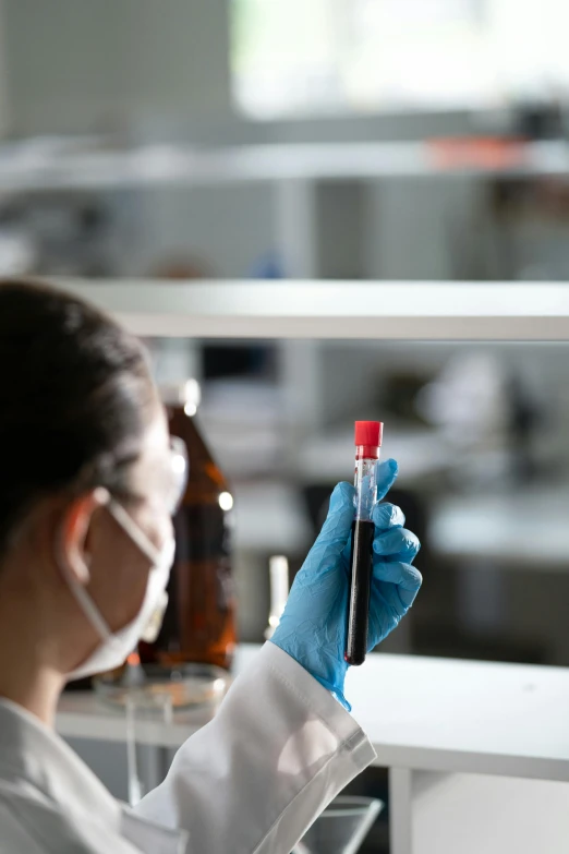 a female in a lab coat and face mask holding a red tube with liquid