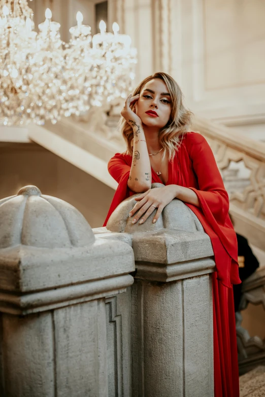 a beautiful blond woman wearing a red dress leaning on a pillar