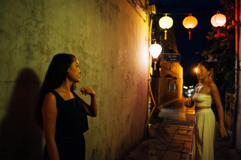 two women stand next to an alley with lanterns above them