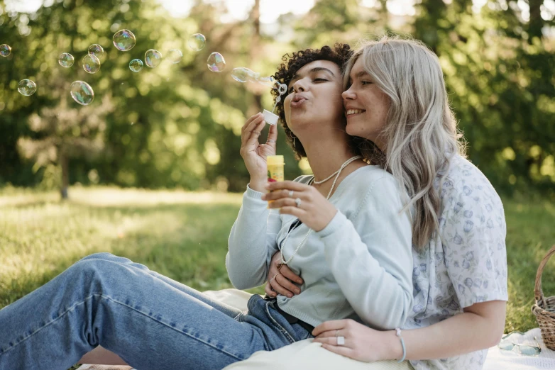 two women with long hair blowing bubbles while sitting on a blanket