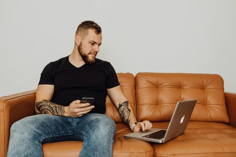 man sitting on brown sofa using cell phone and laptop