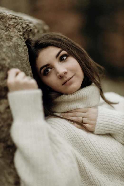 a woman in a white sweater leans against a rock