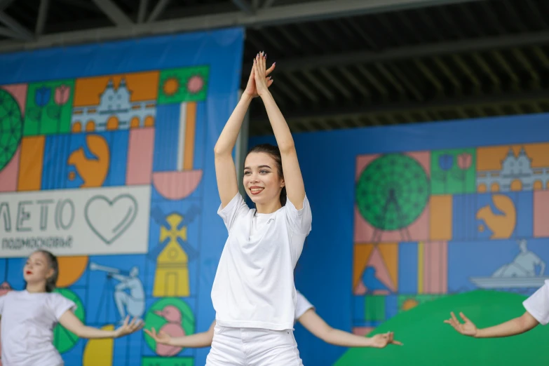 a woman in white clothing stands on stage while other girls are performing