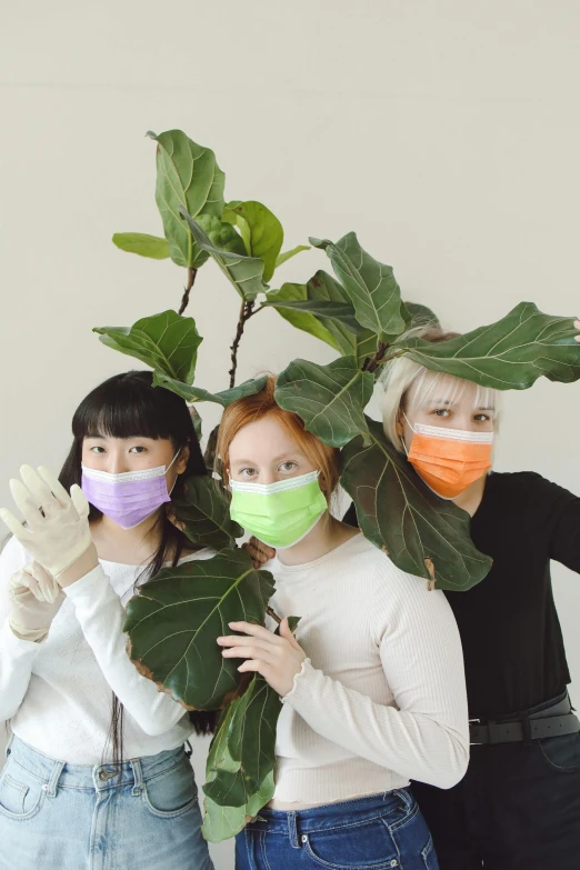 three girls wear face masks while standing behind a plant