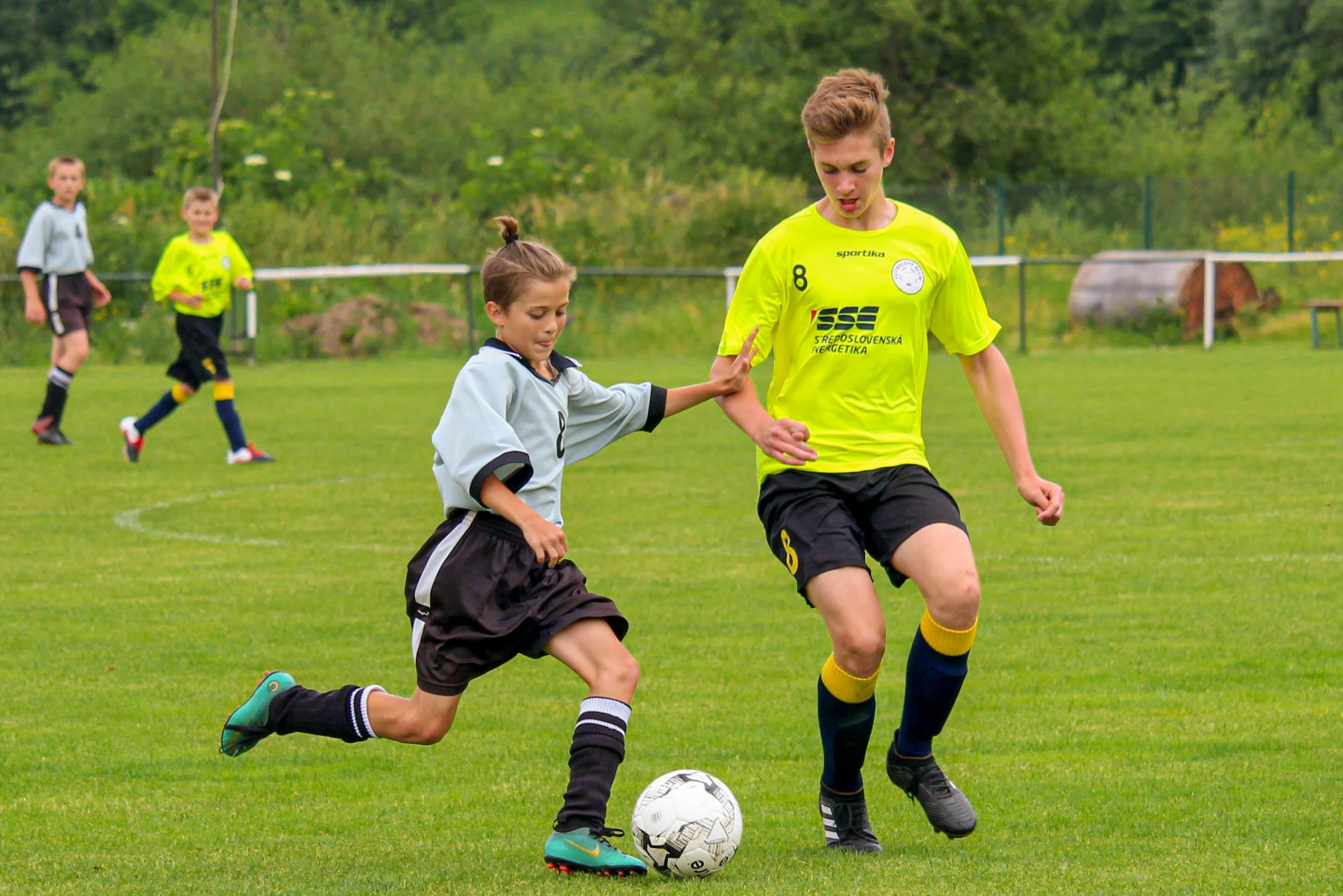 two boys are kicking the soccer ball as others watch