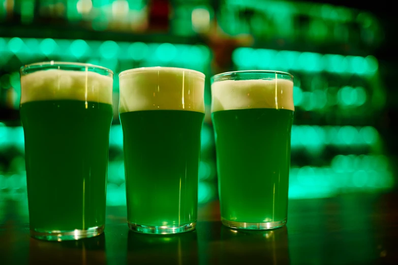 three green drinks sit in front of a row of lit bar lights