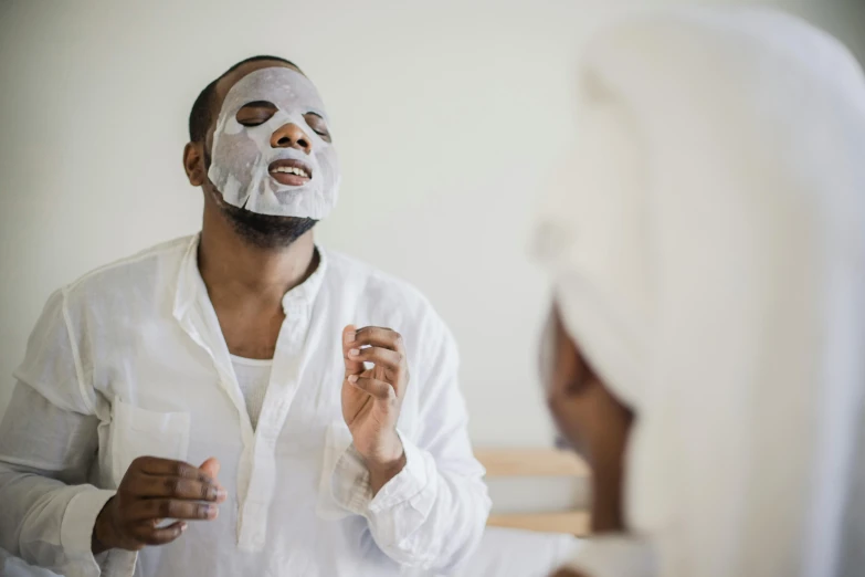 a man wearing a white sheet mask looks at his face in the mirror