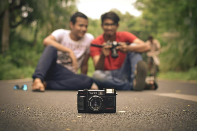 two men sitting on a road next to each other with an old camera in front of them
