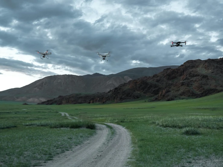 two quad - rotor airplanes flying over the hills and a dirt road