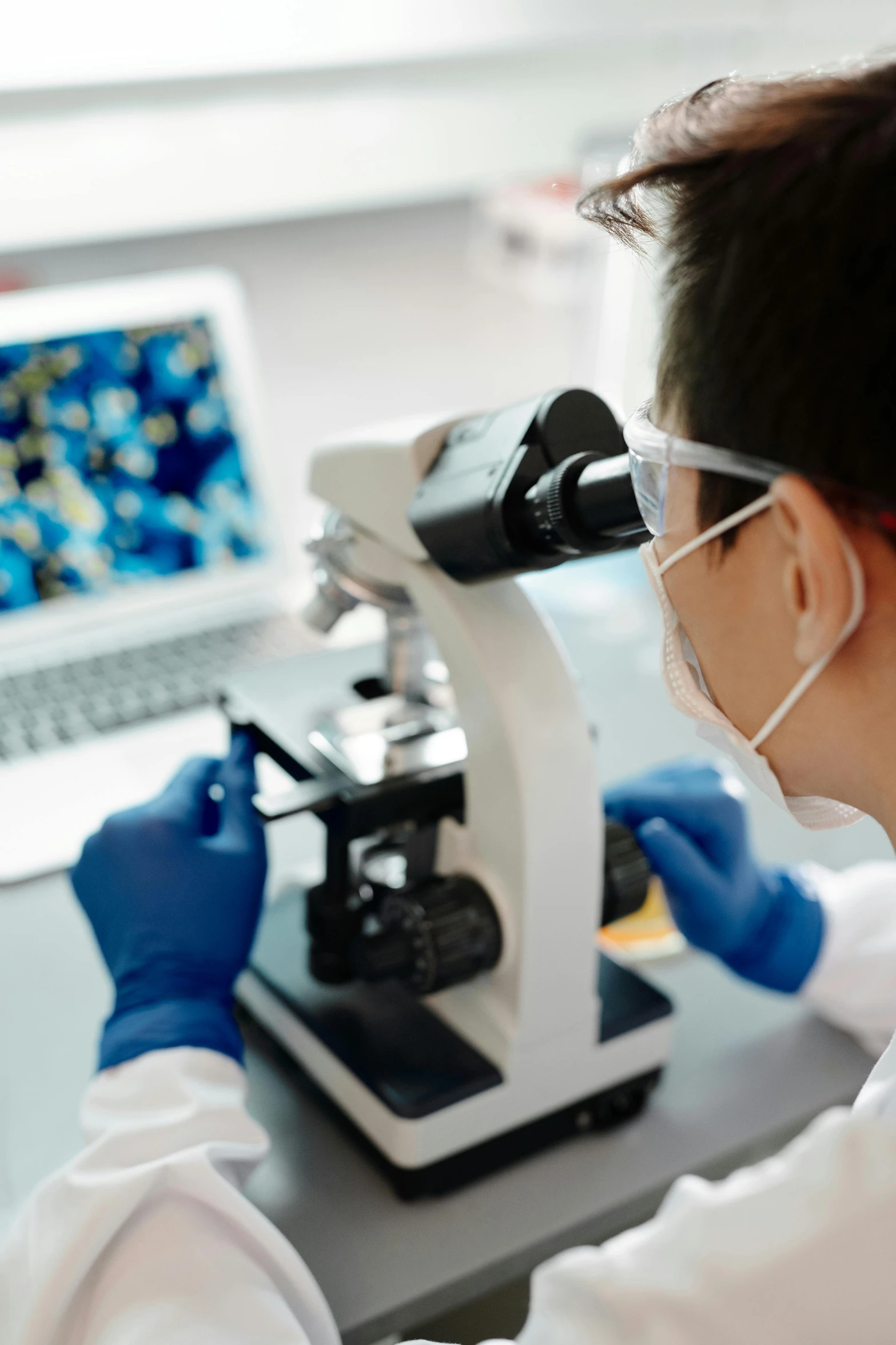 a person in lab coat, glasses and goggles, looking through a microscope