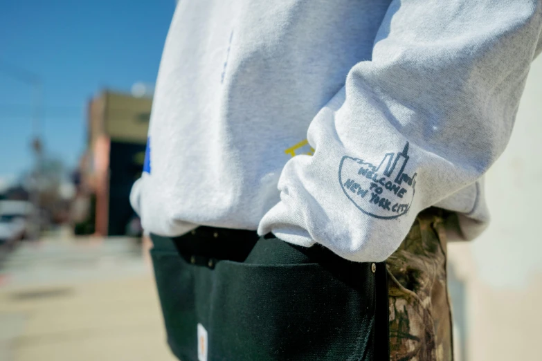 a man's left arm with a sweat shirt on, with a black bottom pocket and two white pants with embroidered logos that have words at the words