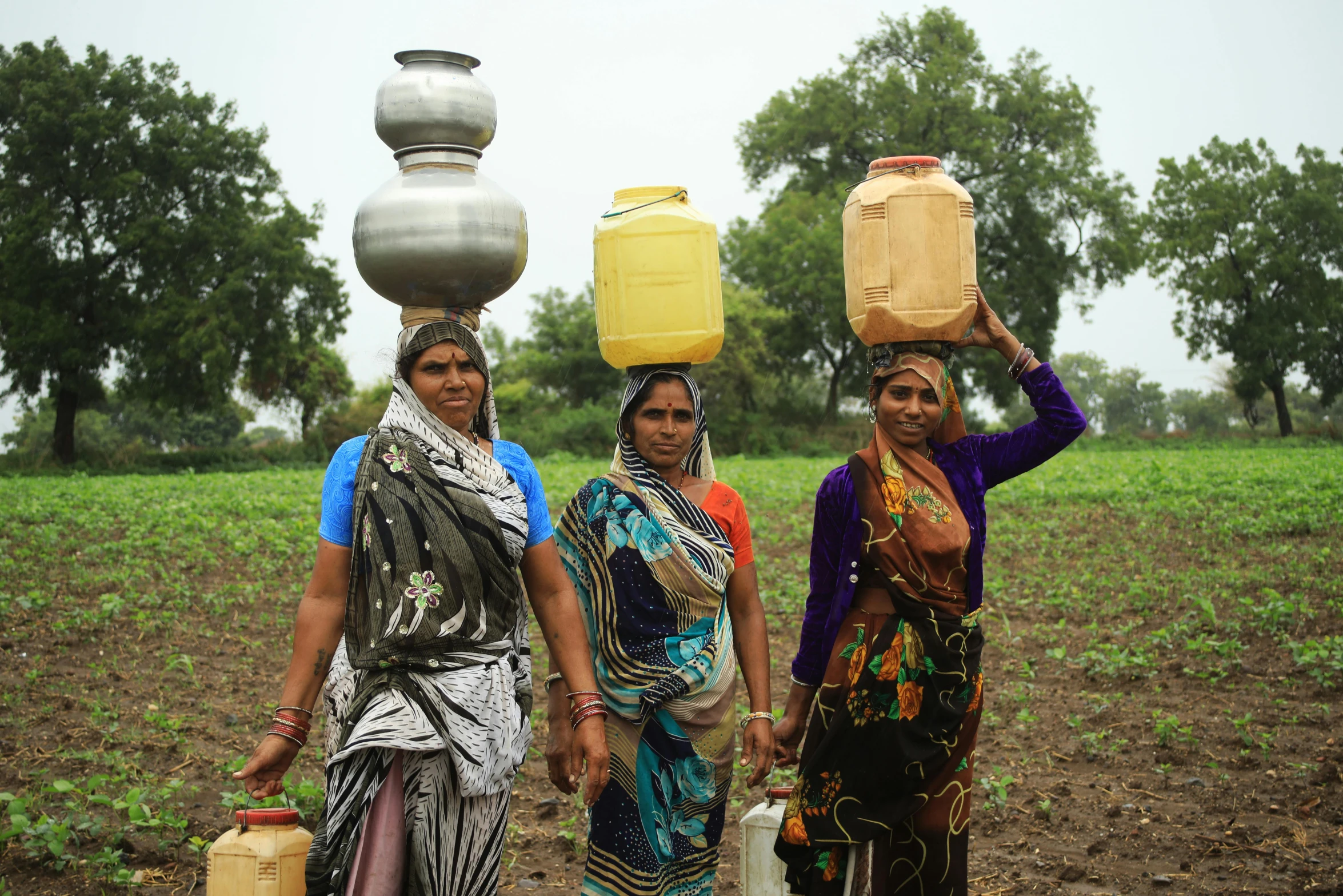 three women walking through a field carrying large water containers