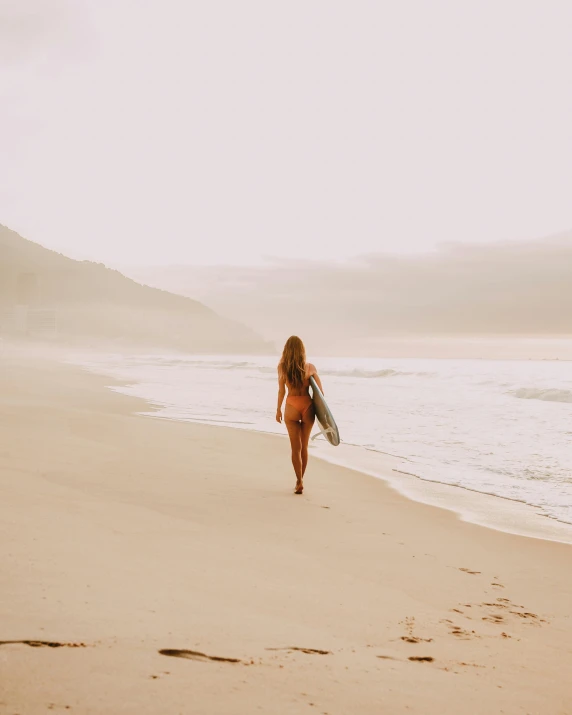 woman walking on beach carrying surfboard with water in the background