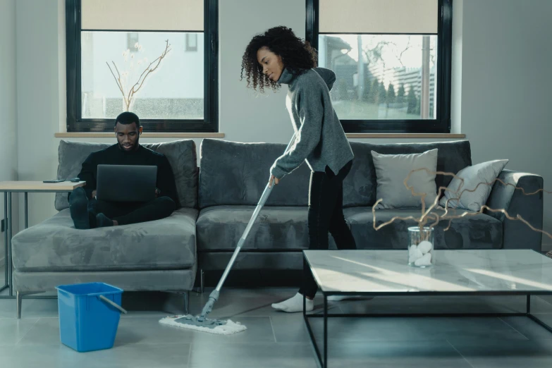 woman is using a vacuum mop as another man uses a laptop computer