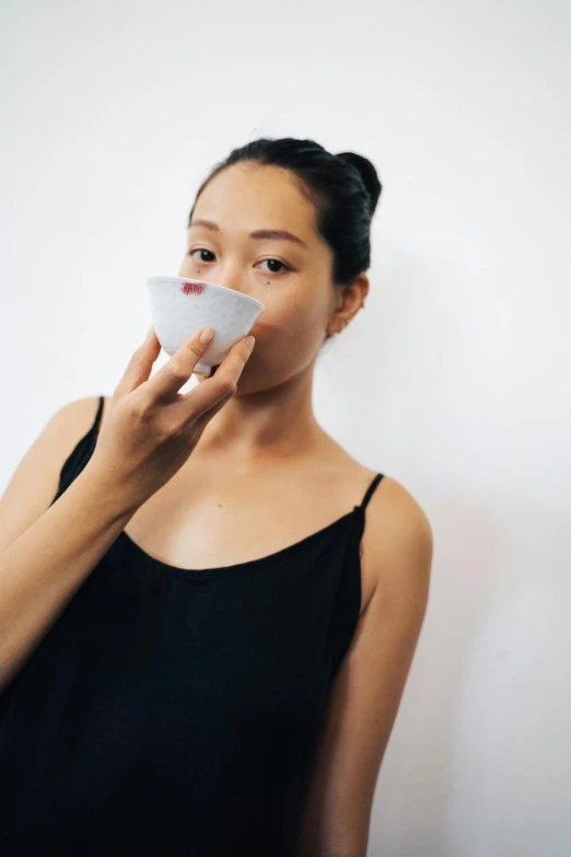a woman standing in front of a white wall holding a cup with her face up to her mouth