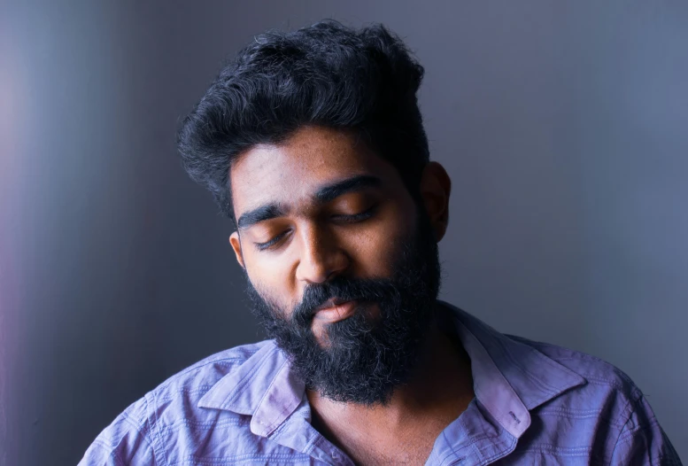 a man with dark hair and a beard wearing a purple shirt looks away