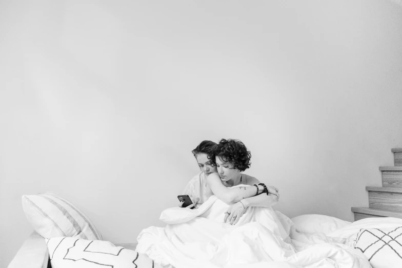 two women in white dresses, one with black hair, the other sitting on a bed and looking at a cell phone