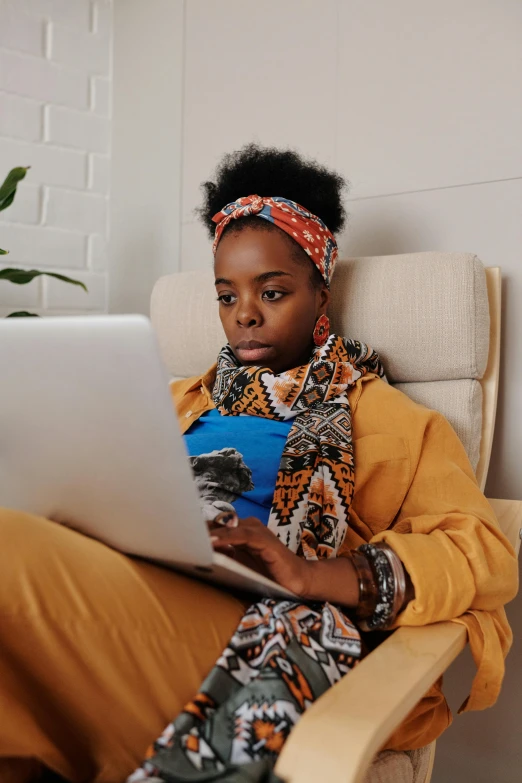 a woman with a bandanna on, sitting in a chair and using a laptop