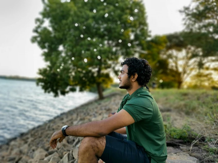 man sitting near water looking at sky with trees