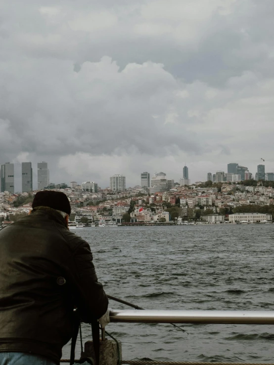 man in black jacket sitting at railing overlooking water and city