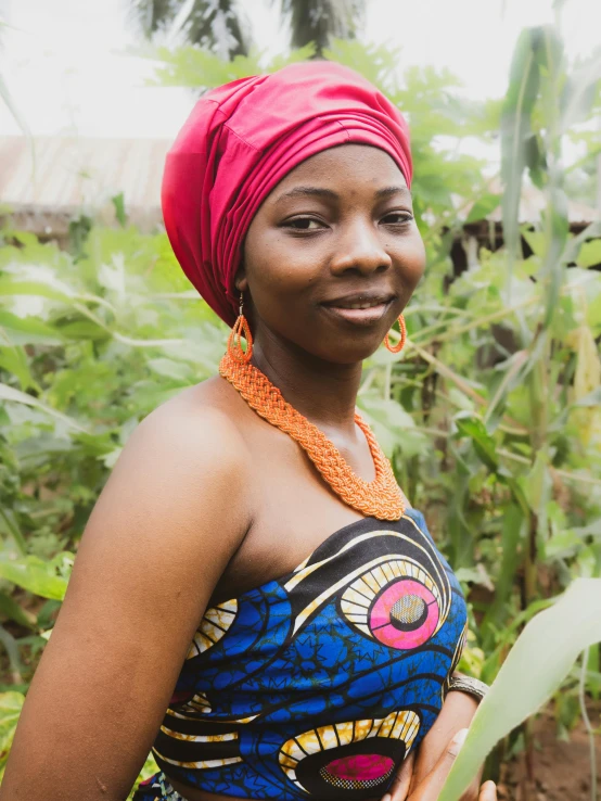 a woman with a red head wrap smiles in front of some plants