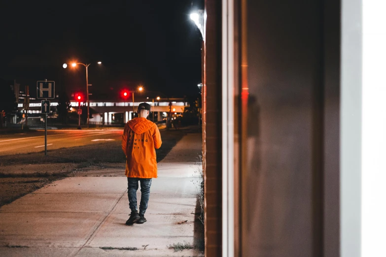 a man in an orange coat is standing on the sidewalk at night