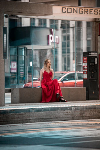 a woman in a red dress sits on a bench at the street curb near a parking meter