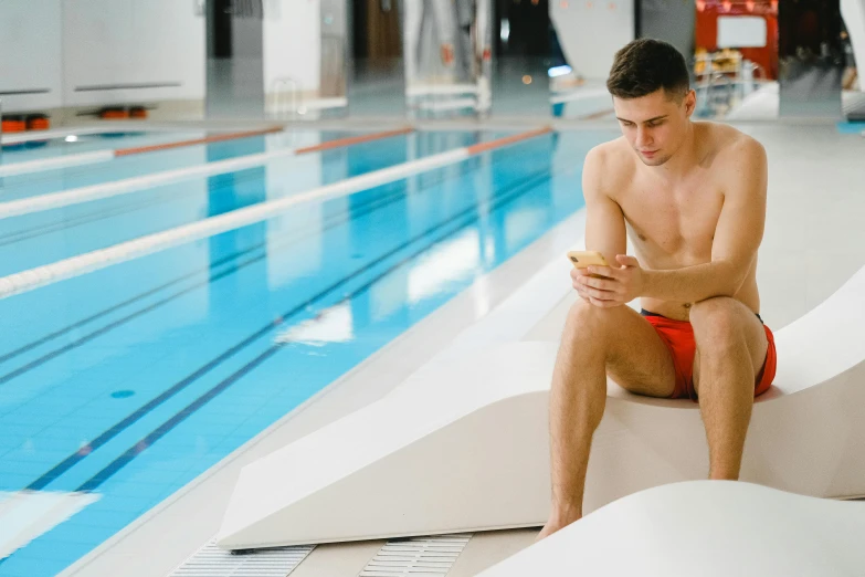 a shirtless man in a red bathing suit is sitting near a pool and looking at his phone