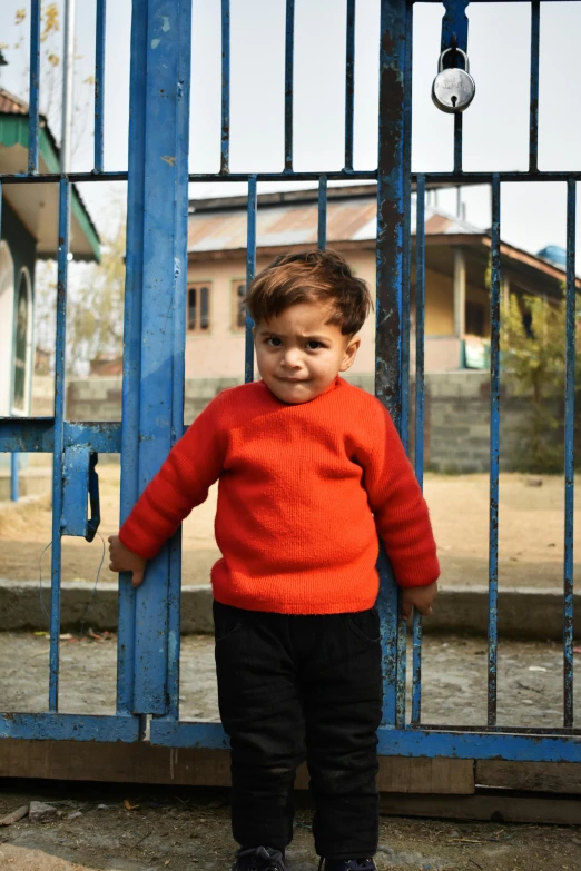 a toddler with red sweater and black pants stands at a blue gate