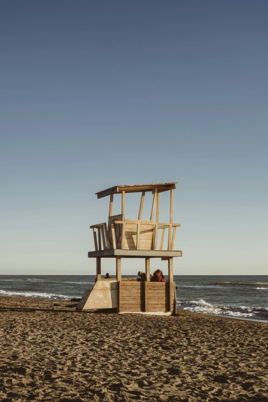 a wooden structure on the beach with a man sitting inside