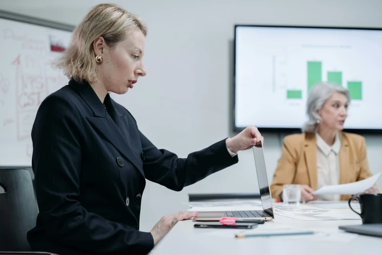 two women sitting at a conference room table with a laptop and other people