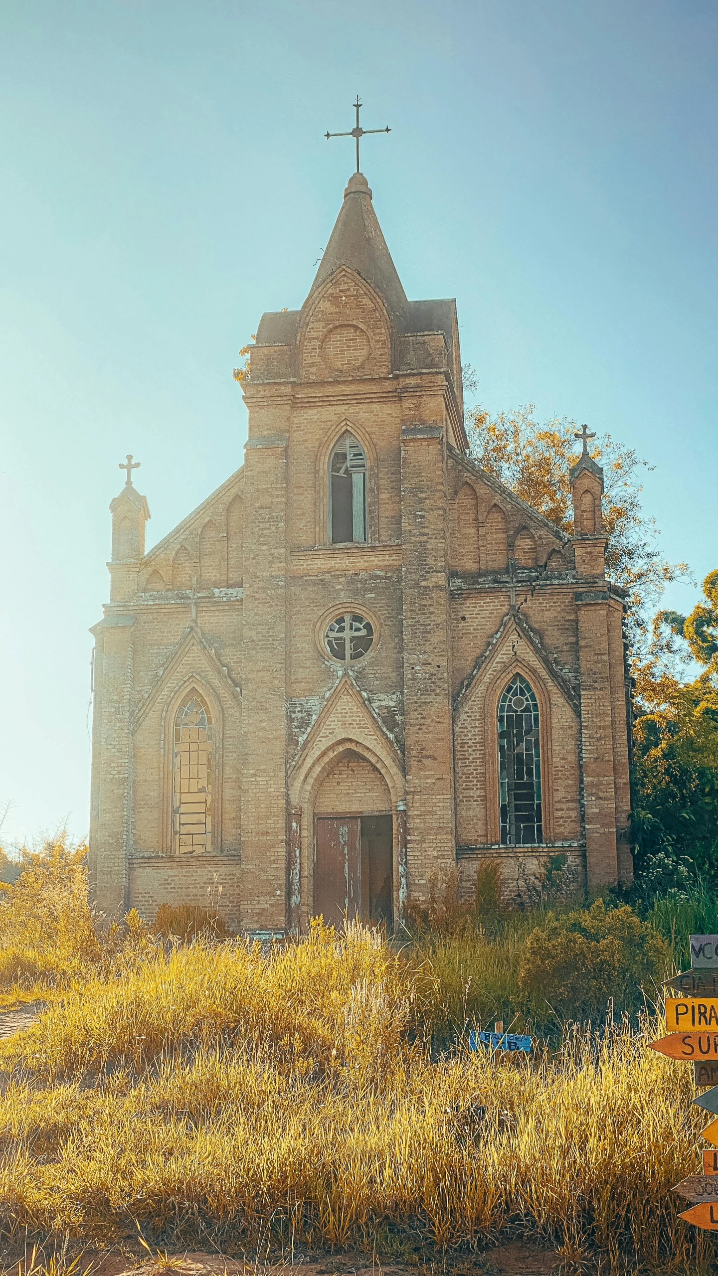 old brick church with cross on top sits on the edge of an overgrown field