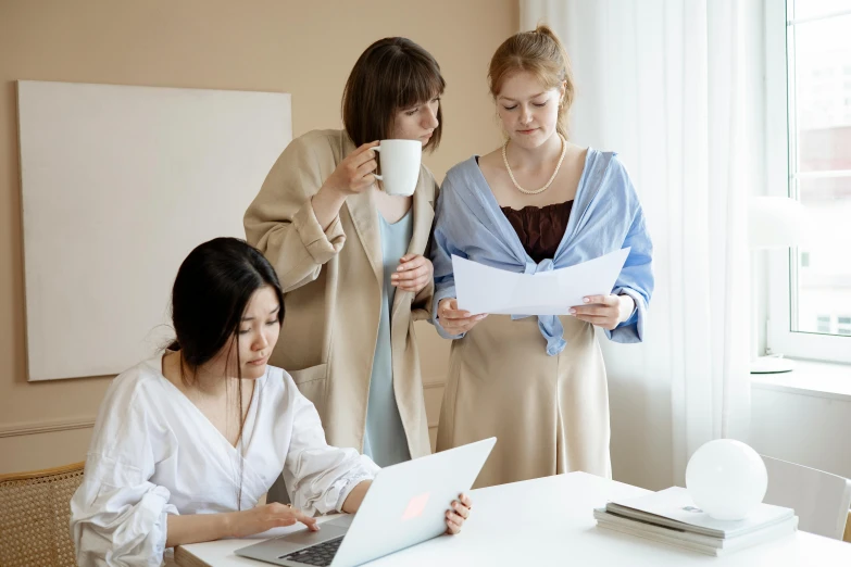 three women work on laptops at a table while looking at a piece of paper