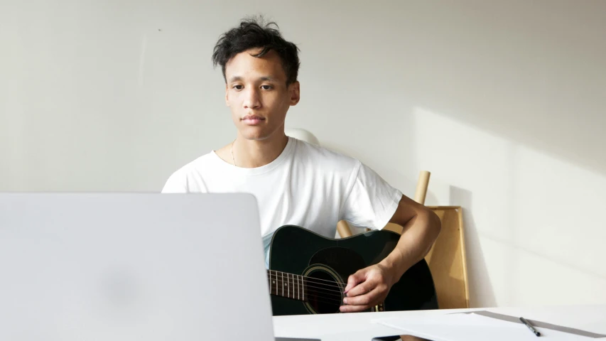 man sitting at table with laptop playing an acoustic guitar