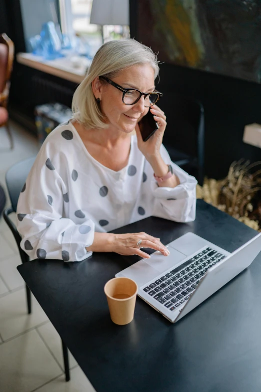 a woman in glasses is talking on her cell phone while sitting at a table with a laptop